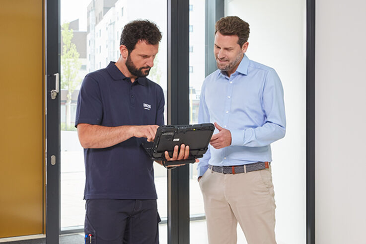 In einem modernen Eingangsbereich zeigt ein Servicemitarbeiter einem Kunden in Business-Outfit technische Informationen auf einem Laptop. 
