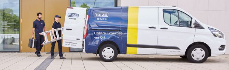 Zwei GEZE Servicemitarbeiter laden Werkzeug und eine Leiter in einen GEZE-Servicewagen vor einem modernen Gebäude.