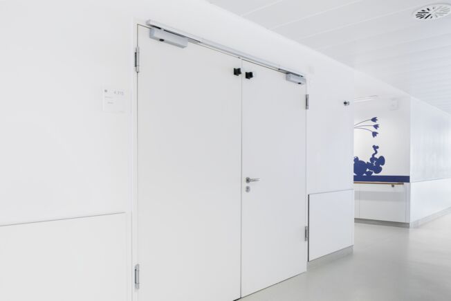 Brandschutztür mit TS 5000 R-ISM und Feststellung über Haftmagnet, verbaut im Olgahospital in Stuttgart. Brandabschnittstür mit TS 5000 R-ISM, Gleitschienentürschließersystem für zweiflügelige Türen mit Schließfolgeregelung und Rauchschalter, Feststellung über Haftmagnet.