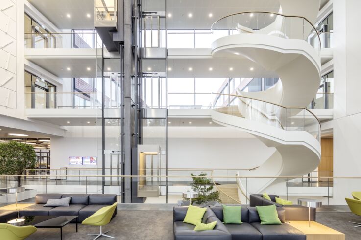 Heller, moderner Eingangsbereich eines Bürokomplexes mit großen Glasflächen, gläsernem Aufzug und Wendeltreppe.