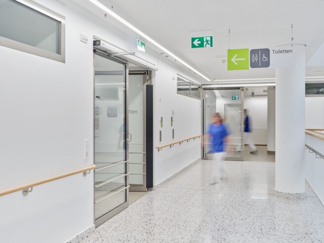 Lys sykehusgang med skyvedører og skilting til toaletter, hvor en pleier går.