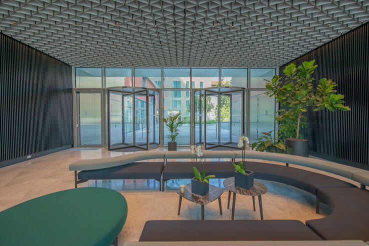 Vista di una hall con divani semicircolari in primo piano, piante verdi e porte girevoli sullo sfondo