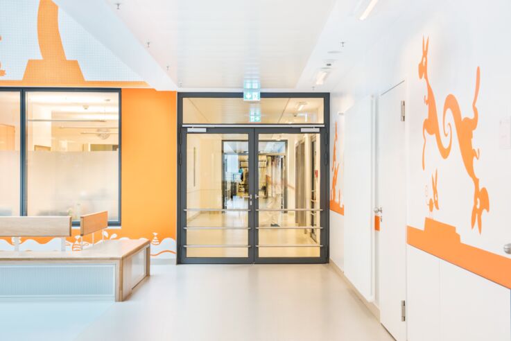 Helle Fluransicht in einem Krankenhaus mit einer großen Glastür, orangen Wandmustern und Sitzbank.